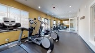 Located at 9107 W Cross Dr in Littleton, Colorado, this fitness center offers a well-appointed, single-floor exercise facility designed to support a comprehensive workout experience. The space is finished with durable rubber flooring and features warm golden-yellow accent walls paired with crisp white trim, creating an energizing yet clean aesthetic throughout.

Recessed lighting illuminates the interior evenly, while multiple windows fitted with roller shades allow ample natural light to fill the room and provide views of the surrounding outdoor greenery. Full-length wall mirrors extend the visual depth of the space and assist users in monitoring their form during exercise.

The cardio zone is well-stocked with multiple treadmills, elliptical trainers, and stationary bikes, all arranged to maximize floor space and usability. A dedicated strength training area includes a multi-function cable machine, a full dumbbell rack with weights of varying sizes, a workout bench, and a selection of medicine balls and accessories. A large wall clock keeps users on schedule during their sessions.

The facility is accessed through a glass-paneled door and also features a set of full-length glass doors opening to the exterior, enhancing ventilation and connectivity to the outdoors. The overall layout is thoughtfully organized, making this fitness center a functional and inviting space for daily exercise.
