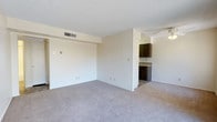This single-floor residence offers a straightforward and functional layout designed for comfortable everyday living. The main living area is generously proportioned, featuring wall-to-wall carpet in a neutral tone that adds warmth underfoot. Smooth, light-colored walls provide a clean backdrop throughout, and a textured ceiling adds subtle visual interest overhead. A ceiling fan is positioned in the living space, contributing to year-round comfort and air circulation. An HVAC vent is integrated into the ceiling, ensuring consistent climate control throughout the space. The living area opens directly toward the kitchen, which is defined by rich, dark wood cabinetry that creates a strong contrast against the lighter surroundings. The kitchen features ample cabinet storage and countertop workspace, making it well-suited for everyday meal preparation. The transition between the carpeted living area and the kitchen is clearly delineated, maintaining a sense of organization within the open layout. Multiple electrical outlets are distributed along the walls, providing practical convenience throughout. The overall ambiance is bright, clean, and move-in ready, offering a neutral canvas that accommodates a wide range of interior styles and furnishing choices.