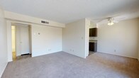 This single-floor residence offers a straightforward and functional layout designed for comfortable everyday living. The main living area is generously proportioned, featuring wall-to-wall carpet in a neutral tone that adds warmth underfoot. Smooth, light-colored walls provide a clean backdrop throughout, and a textured ceiling adds subtle visual interest overhead. A ceiling fan is positioned in the living space, contributing to year-round comfort and air circulation. An HVAC vent is integrated into the ceiling, ensuring consistent climate control throughout the space. The living area opens directly toward the kitchen, which is defined by rich, dark wood cabinetry that creates a strong contrast against the lighter surroundings. The kitchen features ample cabinet storage and countertop workspace, making it well-suited for everyday meal preparation. The transition between the carpeted living area and the kitchen is clearly delineated, maintaining a sense of organization within the open layout. Multiple electrical outlets are distributed along the walls, providing practical convenience throughout. The overall ambiance is bright, clean, and move-in ready, offering a neutral canvas that accommodates a wide range of interior styles and furnishing choices.