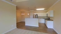 Located at 1850 Popps Ferry Rd in Biloxi, Mississippi, this single-floor residence offers a well-designed open-concept layout that seamlessly connects the kitchen, dining, and living areas. The kitchen is appointed with crisp white cabinetry, granite countertops, stainless steel appliances including a refrigerator and range, and a central island that provides additional prep space and a natural gathering point. Pendant lighting over the island and a flush ceiling fixture enhance both functionality and style. Rich hardwood-style flooring extends throughout the main living areas, adding warmth and continuity to the space. The adjoining dining area is anchored by an elegant chandelier, lending a refined touch to everyday meals. Crown molding trims the ceilings throughout, contributing to the home's polished, cohesive aesthetic. Neutral, warm-toned walls provide a versatile backdrop that complements a range of furnishing styles. Plush carpeting in the adjacent living area offers a comfortable contrast to the hard flooring. The overall ambiance is bright, clean, and welcoming, making this single-story home a practical yet stylish residence in the Biloxi community.