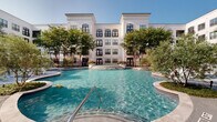 This beautifully designed outdoor amenity space serves as the centerpiece of a sophisticated multi-story residential complex, characterized by a classic white façade, arched windows, and dark-trimmed detailing that lends the architecture a refined, timeless aesthetic. The focal point of the space is a generously sized freeform swimming pool with shimmering turquoise water, featuring a gradual entry point and decorative water features along its curved edges. The pool is framed by a spacious stone-finished sundeck furnished with lounge chairs, umbrella-shaded seating areas, and stylish patio furniture — ideal for relaxation and social gatherings. Mature trees are artfully integrated throughout the courtyard, including island planters within the pool deck that provide natural shade and a lush, park-like atmosphere. A separate green lawn area is visible from the pool terrace, adding to the resort-quality feel of the complex. The space is accessed through a secured ornamental iron gate, enhancing both privacy and curb appeal. Surrounding balconies on the multi-story building overlook the courtyard, creating a sense of community while offering residents elevated views of this vibrant outdoor retreat. The overall ambiance is bright, open, and elegantly appointed.