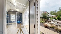 This single-story community amenity space combines an indoor fitness center with a resort-style outdoor pool area, offering residents a comprehensive recreational experience. The fitness center is thoughtfully designed with vaulted ceilings, light hardwood-style flooring, and ceiling fans that promote airflow throughout the space. Large wall mirrors extend the sense of space visually, while natural light filters in through multiple windows. The room is equipped with a variety of cardio machines, including treadmills and cycling bikes, as well as a dedicated strength-training area featuring dumbbells, resistance equipment, and foam rollers neatly organized on wall-mounted racks. A bold dark accent wall adds a dynamic, modern aesthetic to the interior. Access to the fitness center is through glass-paneled doors clearly labeled "Fitness Center," opening to a well-maintained outdoor walkway. The outdoor pool area is equally impressive, featuring a generously sized rectangular swimming pool with clear turquoise water, surrounded by a spacious concrete deck lined with lounge chairs for sunbathing. Two pergola structures with seating areas offer shaded retreats, while lush mature trees provide a natural, private backdrop. The overall environment is serene, well-maintained, and designed to support an active and relaxed lifestyle.