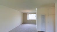 This single-story space presents a clean, understated interior designed with simplicity and functionality in mind. The main living area is finished with wall-to-wall carpeting in a neutral gray tone, contributing to a quiet, comfortable atmosphere underfoot. Warm beige walls are complemented by white crown molding along the ceiling, adding a subtle architectural detail that frames the room neatly.

A centrally hung pendant light fixture serves as a modest focal point, providing ambient illumination throughout the space. Natural light enters through a large window along the far wall, fitted with vertical blinds that allow for adjustable light control while offering views to the outdoors. The window brightens the interior considerably, enhancing the open feel of the room.

On the right side of the space, a louvered panel — likely concealing a utility or HVAC unit — adds a practical element to the layout. The smooth, light-colored ceiling further reflects natural light, keeping the interior feeling airy. Overall, the space maintains a neutral, move-in-ready aesthetic with a calm and functional character throughout.