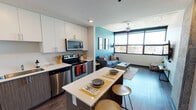 This contemporary residential unit is thoughtfully designed with a seamless open-concept layout connecting the kitchen, dining, and living areas. The kitchen is well-appointed with stainless steel appliances — including a refrigerator, electric range, microwave, and dishwasher — complemented by two-tone cabinetry featuring crisp white upper cabinets and warm wood-grain lower cabinets, all paired with clean white countertops. A central kitchen island with bar seating provides additional prep and dining space. The living area makes a bold statement with a vibrant teal accent wall, exposed concrete ceiling, dark hardwood-style flooring, and a ceiling fan. Floor-to-ceiling windows flood the space with natural light and frame open urban views. The bedrooms are carpeted and feature large windows with urban outlooks, built-in storage including sliding closet doors, and dedicated desk areas suited for study or work. The bathroom is finished with clean tile flooring, a wood-grain vanity with white countertop, an oversized mirror, and bright overhead lighting. Throughout the unit, the industrial character of the exposed concrete ceilings is balanced by modern, clean-lined finishes, creating a vibrant and functional living environment.