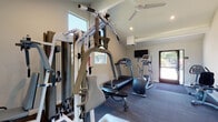 Located at 555 Elmira Rd in Vacaville, California, this fitness center offers a well-equipped, single-floor workout facility designed to support a comprehensive exercise routine. The space features vaulted ceilings that lend an open, airy feel, while large windows along the walls flood the interior with abundant natural light and provide pleasant views of the surrounding mature trees and landscaping outside.

The fitness center is divided into distinct functional zones. One area houses a multi-station cable weight machine alongside a flat bench, providing ample options for strength training and resistance exercises. Adjacent to this, a dedicated rack of hex dumbbells in varying weights offers versatility for free-weight workouts, complemented by a large wall-mounted mirror that helps users monitor their form.

The cardio section is well-appointed with a treadmill, an elliptical trainer, and an upright stationary bike, all positioned near a wall-mounted flat-screen television for entertainment during workouts. A ceiling fan overhead helps maintain comfortable airflow throughout the space. The floors are finished with durable, dark carpet tiles suitable for a fitness environment. Natural light pours in through multiple windows, and direct exterior access via a glass-paneled door adds convenience. The overall ambiance is clean, modern, and functional.