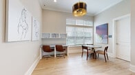 Located at 2150 NW 21st St in Ocala, Florida, this single-floor professional space offers a refined and functional environment designed to support both focused work and collaborative meetings. The interior is finished with light hardwood-style flooring and neutral warm-toned walls, creating a clean and inviting atmosphere throughout.

The main area features a built-in L-shaped desk workstation with open white wall-mounted shelving, accommodating multiple users with comfortable leather rolling chairs. A large window fitted with horizontal blinds floods the workspace with generous natural light while offering views of the surrounding greenery. A round pedestal conference table with upholstered seating provides an ideal spot for small group discussions or informal meetings.

Overhead, a striking cylindrical drum pendant light with a gold interior serves as a bold focal point, adding a touch of contemporary elegance. The space is adorned with curated artwork, including an equestrian line drawing and a vibrant abstract canvas in rich reds and purples, lending a sophisticated, gallery-like quality to the environment.

A notable design feature is a textured reclaimed wood accent wall visible through a glass-paned interior door, adding warmth and architectural interest. Crown molding and clean white trim throughout lend a polished finish. The layout flows openly, making efficient use of the available space while maintaining an uncluttered, professional ambiance.