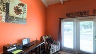 This single-floor space makes an immediate impression with its bold, warm orange walls that create an energetic and inviting atmosphere. The room draws the eye upward to its charming white-painted exposed beam ceiling, which adds architectural character and a sense of height to the interior.

Natural light filters in through a window dressed with dark curtain panels and horizontal blinds, offering a view of the lush greenery outside. A decorative "WELCOME" sign above the window reinforces the space's warm and hospitable tone.

The layout thoughtfully balances productivity and relaxation. A sleek, dark-toned desk accompanied by a matching chair serves as a dedicated workspace, while a stylish patterned armchair with a bold polka-dot design provides a comfortable spot to unwind. A piece of artwork on the wall adds a personal, curated touch to the overall aesthetic.

The flooring transitions from hard surface to a textured area rug, grounding the seating area and adding warmth underfoot. Together, the architectural details, vibrant color palette, and carefully chosen furnishings give this space a distinctive personality — equal parts functional home office and cozy retreat.
