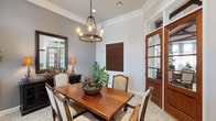 This single-story space showcases a well-appointed dining room defined by classic architectural details and warm, traditional furnishings. Crown molding lines the ceiling throughout, adding a polished finishing touch to the soft gray walls. A wrought-iron candle-style chandelier serves as a central focal point, suspended above a solid wood dining table surrounded by upholstered chairs with carved wooden frames, all set atop a decorative patterned area rug.

Natural light enters through multiple windows dressed with striped gray and cream panel curtains paired with wood-toned blinds, offering views of an outdoor patio area. A dark wood credenza along one wall features ornate cabinet doors and is styled with matching table lamps and a large framed mirror, enhancing the room's depth and elegance. Potted tropical plants are placed throughout, introducing organic texture and color to the space.

A glass-paneled interior door with rich wood framing connects to an adjacent room, contributing to an open and cohesive flow. The overall ambiance is warm and inviting, blending traditional design elements with refined detailing to create a comfortable and stylish dining environment.