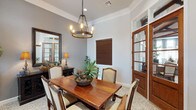 This single-story space showcases a well-appointed dining room defined by classic architectural details and warm, traditional furnishings. Crown molding lines the ceiling throughout, adding a polished finishing touch to the soft gray walls. A wrought-iron candle-style chandelier serves as a central focal point, suspended above a solid wood dining table surrounded by upholstered chairs with carved wooden frames, all set atop a decorative patterned area rug.

Natural light enters through multiple windows dressed with striped gray and cream panel curtains paired with wood-toned blinds, offering views of an outdoor patio area. A dark wood credenza along one wall features ornate cabinet doors and is styled with matching table lamps and a large framed mirror, enhancing the room's depth and elegance. Potted tropical plants are placed throughout, introducing organic texture and color to the space.

A glass-paneled interior door with rich wood framing connects to an adjacent room, contributing to an open and cohesive flow. The overall ambiance is warm and inviting, blending traditional design elements with refined detailing to create a comfortable and stylish dining environment.