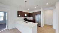 This single-floor space showcases a thoughtfully designed, open-concept kitchen that serves as a central highlight. The kitchen features rich, dark wood cabinetry that contrasts elegantly against crisp white quartz countertops, creating a bold yet sophisticated aesthetic. A generously sized kitchen island with an integrated double sink anchors the space, offering both functional workspace and a natural gathering point.

Two cylindrical pendant lights hang above the island, providing focused task lighting while adding a modern decorative touch. Recessed ceiling lights further illuminate the area, ensuring a bright and welcoming atmosphere throughout. Stainless steel appliances, including a full-sized refrigerator, gas range with microwave above, and built-in dishwasher, lend a professional-grade quality to the cooking space.

Upper and lower cabinetry with sleek hardware provides ample storage, while the light wood-tone flooring throughout adds warmth and continuity. White walls and a clean, minimalist design sensibility keep the space feeling open, airy, and contemporary. A hallway visible in the background suggests additional rooms beyond the main kitchen area.
