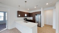 This single-floor space showcases a thoughtfully designed, open-concept kitchen that serves as a central highlight. The kitchen features rich, dark wood cabinetry that contrasts elegantly against crisp white quartz countertops, creating a bold yet sophisticated aesthetic. A generously sized kitchen island with an integrated double sink anchors the space, offering both functional workspace and a natural gathering point.

Two cylindrical pendant lights hang above the island, providing focused task lighting while adding a modern decorative touch. Recessed ceiling lights further illuminate the area, ensuring a bright and welcoming atmosphere throughout. Stainless steel appliances, including a full-sized refrigerator, gas range with microwave above, and built-in dishwasher, lend a professional-grade quality to the cooking space.

Upper and lower cabinetry with sleek hardware provides ample storage, while the light wood-tone flooring throughout adds warmth and continuity. White walls and a clean, minimalist design sensibility keep the space feeling open, airy, and contemporary. A hallway visible in the background suggests additional rooms beyond the main kitchen area.