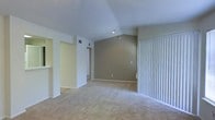 This single-story space showcases a thoughtfully designed open layout with notable architectural interest provided by vaulted ceilings that lend a sense of spaciousness and airiness to the main living area. The interior is finished in a neutral color palette, combining soft white and warm taupe wall tones that create a calm, welcoming ambiance throughout.

The living area is anchored by plush wall-to-wall carpeting in a light beige tone, contributing to a comfortable and quiet atmosphere underfoot. A large sliding glass door fitted with white vertical blinds lines one wall, allowing for generous natural light while offering privacy and direct access to the outdoors.

Recessed lighting in the ceiling supplements natural illumination, ensuring the space remains well-lit. Clearly defined transitional areas branch off from the main room, with open passageways leading to additional rooms and hallways, suggesting a functional and connected floor plan. White structural columns and clean architectural lines further define the space without disrupting the open feel.

Overall, the space presents a clean, move-in ready environment with a neutral aesthetic well-suited for a variety of interior styles.