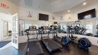 Located at 45 Farm Rd in Hillsborough, NJ, this single-story amenity facility is designed to support both fitness and everyday convenience. The fitness area is the centerpiece of the space, featuring elevated ceilings that create an open, airy atmosphere. Large wall-mounted mirrors visually expand the room, while recessed and overhead lighting ensure a bright workout environment. The cardio zone is equipped with multiple treadmills and elliptical machines, complemented by a wall-mounted flat-screen TV for entertainment during workouts. A separate strength training area includes upright stationary bikes, adjustable weight benches, a cable multi-gym machine, and a fully stocked dumbbell rack, providing a well-rounded selection of equipment. Motivational fitness-themed wall art and decals contribute to an energizing ambiance throughout. Dark flooring throughout the fitness rooms provides a clean, modern aesthetic. Adjacent to the fitness area, the laundry room features multiple stacked washer and dryer units arranged along the walls, with speckled tile flooring that is easy to maintain. A small common area with a wall-mounted TV, folding table, and seating offers a convenient waiting or gathering space, rounding out this practical and functional facility.