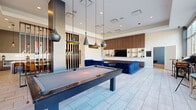 Located at 998 Church St in Glenview, Illinois, this single-floor amenity space offers a well-appointed, contemporary environment designed for socializing and entertainment. The open-concept layout is anchored by large floor-to-ceiling windows that flood the interior with natural light and provide expansive views of the surrounding streetscape.

The main lounge area is a standout feature, showcasing bold royal blue tufted velvet sectional sofas arranged around a sleek marble-top coffee table. A full-wall built-in media center, finished in warm walnut tones with open display shelving and a mounted flat-screen TV, serves as the focal point of the lounge. A billiards table positioned beneath a dramatic cluster of modern pendant lights adds a recreational element to the space.

The kitchen is thoughtfully designed with dark walnut cabinetry, crisp white quartz countertops, and a generous center island with bar seating for multiple guests. Stainless steel appliances, including a beverage refrigerator, complement the contemporary aesthetic. Mixed metallic and glass pendant lights illuminate the workspace, while vibrant artwork adds a pop of color.

Light-toned large-format tile flooring flows throughout the entire space, unifying each zone seamlessly. The entry features floor-to-ceiling glazing, reinforcing the bright and welcoming atmosphere of this well-curated communal space.