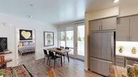 This single-floor residence offers a thoughtfully designed layout with a clean, modern aesthetic defined by white walls, light wood-style flooring, and an abundance of natural light. The open-plan living and dining area forms the heart of the home, where a comfortable seating area with a gray sofa, colorful area rug, and media shelving flows seamlessly into a dedicated dining space. The dining area is anchored by a solid wood table and is positioned beside full-length glass doors that open to a private outdoor balcony, framing views of surrounding greenery.

The kitchen is well-equipped with gray cabinetry, white subway tile backsplash, under-cabinet lighting, and a full suite of stainless steel appliances including a refrigerator, range, microwave, and dishwasher. A freestanding wood island provides additional prep and storage space.

Two bedrooms offer comfortable retreats; one features a glass door with direct outdoor access and a side window that floods the room with natural light. The bathroom includes a vanity with gray cabinetry, a white countertop, a full bathtub with shower, and recessed lighting. Adjacent closet space adds practical storage. Overall, the residence presents a cohesive, contemporary living environment.