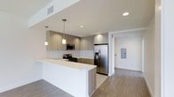 This single-floor space showcases a thoughtfully designed open-concept kitchen that seamlessly connects to adjoining living areas. The kitchen is anchored by a generous island with a crisp white countertop, providing ample workspace and a natural gathering point. Flat-panel cabinetry in a warm wood-grain finish lines the walls, complemented by a classic white subway tile backsplash that adds texture while maintaining a clean aesthetic.

Stainless steel appliances, including a French door refrigerator with an in-door water and ice dispenser, a gas range, and an over-the-range microwave, deliver a polished, professional feel. Recessed ceiling lighting is supplemented by two stylish pendant lights suspended above the island, creating layered illumination throughout the kitchen.

The flooring transitions from light-toned tile within the kitchen to warm hardwood-style planks in the adjacent areas, subtly defining each zone. Neutral white walls and ceilings enhance the sense of openness and brightness throughout. An electrical panel is discreetly positioned along one wall of the adjacent space, maintaining the clean, uncluttered character of the overall design.