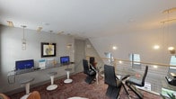 Located in Spring, TX, this contemporary upper-level space is thoughtfully designed to serve as a functional and stylish workspace or business lounge. The room features patterned carpet in warm red and gray tones that grounds the space and adds visual texture throughout.

A dedicated computer workstation area lines one wall, equipped with a sleek glass-top desk, multiple computer terminals, and modern pendant lighting with gold accents suspended from the ceiling. Cushioned pedestal stools provide casual seating at the workstation, complemented by a retro-inspired accent chair in a colorful striped fabric. Framed artwork in yellow, black, and white tones adorns the walls, adding an artistic element to the professional setting.

At the center of the room sits a round glass dining or conference table surrounded by contemporary black chairs with chrome frames, ideal for small group meetings or collaborative work sessions. A large-scale abstract canvas painting serves as a striking focal point on one wall, while potted plants introduce a touch of greenery throughout the space.

Natural light filters in through windows fitted with white blinds, and an open railing with a wood-and-cable design borders one side of the room, offering a view to the level below. The angled ceiling adds architectural interest and a sense of dynamic volume to the overall design.