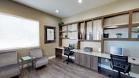 This single-story space is designed to function as a dedicated workspace, combining functionality with a clean, contemporary aesthetic. The focal point of the room is a built-in workstation featuring a continuous light-colored countertop that spans the width of the back wall, paired with lower cabinetry in a warm walnut-toned finish. Above the desk, an open shelving unit rises to the ceiling, offering generous display and storage space organized into a grid-like configuration. The shelves are currently accented with decorative vases and greenery, adding visual interest to the otherwise streamlined design.

The flooring consists of dark hardwood-style planks that contrast effectively against the warm beige walls, grounding the space with a sense of warmth and durability. Recessed ceiling lights provide consistent illumination throughout the room, complemented by natural light filtering in through a window fitted with horizontal blinds on the left wall. A framed botanical print adds a subtle decorative touch beside the window. A cushioned accent chair is positioned nearby, offering a secondary seating area. Overall, the space conveys a practical yet polished environment well-suited for office or administrative use.