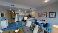 This single-floor space showcases an open-concept layout that seamlessly connects the kitchen, dining, and living areas into one cohesive environment. The kitchen is outfitted with white shaker-style cabinetry, granite countertops, and stainless steel appliances, including a full-size refrigerator, microwave, and dishwasher. A peninsula with bar seating provides a casual dining option and acts as a natural divider between the kitchen and the rest of the space. Recessed lighting and flush-mount ceiling fixtures illuminate the kitchen and living areas effectively. The flooring throughout features a striking polished concrete in a deep blue-grey tone, adding an industrial yet modern character to the space. The dining area accommodates a round white table paired with navy blue upholstered chairs, creating an intimate setting. The living area features a grey sofa accented with decorative throw pillows, anchored by a woven area rug. Wall art and floral arrangements add warmth and personality to the space. Overall, the design balances contemporary aesthetics with functional living in a well-organized, open floor plan.