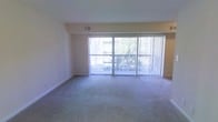 This single-floor space features a spacious living area finished with wall-to-wall carpeting in a neutral tone, contributing to a clean and understated aesthetic. The room is anchored by a large sliding glass door framed by vertical blinds, which spans much of the exterior wall and opens onto a private balcony with a metal railing. This expansive glazing allows abundant natural light to fill the interior, creating an airy and open atmosphere. Glimpses of mature trees are visible beyond the balcony, providing a touch of greenery to the outdoor view. The walls are finished in a warm neutral tone, and the ceiling is smooth with a standard height that maintains a comfortable sense of scale throughout the room. A ceiling vent is visible, indicating a central HVAC system for climate control. The baseboards are clean and white, neatly framing the carpeted floor. The overall ambiance of the space is calm and versatile, offering a blank canvas well-suited for a variety of furniture arrangements and interior styles.