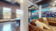 This single-floor community lounge showcases a striking industrial aesthetic defined by exposed brick walls, polished concrete floors, and exposed ductwork running along the dark-painted ceiling. The open layout is anchored by a bold, colorful mural depicting a mountain landscape with the word "Ogden" rendered in large letters, serving as a vivid focal point that celebrates the local surroundings. The space is divided into distinct activity zones that flow seamlessly into one another. A comfortable lounge area features tufted leather sofas, patterned area rugs, and a mounted flat-screen television set against the warm texture of the brick wall. A dedicated gaming area includes a full-size ping-pong table, a foosball table, and an arcade game, providing ample entertainment options. A dining and social zone offers high-top tables and stools positioned in front of the mural, ideal for casual gatherings. Large windows along the perimeter flood the space with natural light and offer views of the surrounding greenery, enhancing the open, airy atmosphere. Structural columns punctuate the floor plan, adding to the loft-like character of the space. Potted plants and carefully chosen décor elements soften the industrial materials, creating an energetic yet welcoming environment suited for socializing and recreation.