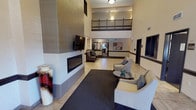 This property makes a strong first impression with its grand lobby, characterized by soaring double-height ceilings and an open mezzanine level overlooking the main floor. The lobby is designed for comfort and socializing, anchored by a sleek linear gas fireplace set into a large tile-clad feature wall, above which a flat-screen television is mounted. Stylish lounge seating, including upholstered armchairs arranged around a glass-topped coffee table, invites guests to relax on a large area rug over tile flooring. Warm, neutral tones and accent lighting create an inviting ambiance throughout.

The fitness center is a well-appointed, dedicated space designed to support a full workout routine. Dark rubberized flooring lines the room, which is equipped with commercial-grade cardio machines including treadmills and an elliptical trainer from TRUE Fitness, as well as a multi-function Paramount cable strength machine and free weights. A full-length mirror spans one wall, enhancing the sense of space and allowing users to monitor their form. Natural light enters through horizontal-blind-fitted windows, while overhead fluorescent lighting ensures the room is well-lit. A wall-mounted television, exercise balls, and a conveniently placed hydration station with a water fountain, sanitation supplies, and paper towel dispenser round out the amenities.