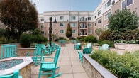 Located at 1 Glover Ave in Norwalk, Connecticut, this well-appointed outdoor courtyard serves as a central gathering space within a multi-story residential building. The patio is finished with large-format tile pavers that provide a clean, durable surface throughout the space. A standout feature is the octagonal fire pit, framed in stone with blue glass accents, and surrounded by vibrant teal Adirondack-style rocking chairs that lend a relaxed, welcoming atmosphere. Additional seating areas with tables and chairs are positioned throughout the courtyard, offering a variety of spaces for both social gatherings and quiet relaxation. The courtyard is bordered by raised stone planters with smooth coping details, filled with an abundance of carefully maintained, diverse plantings including ornamental shrubs, flowering perennials, and mature trees that provide privacy and greenery throughout the seasons. Classic black lamp posts add architectural interest and ensure the space is well-lit. The surrounding multi-story building with balconies wraps around the courtyard, creating an enclosed, sheltered feel while still allowing ample natural light. The overall ambiance is inviting and resort-like, designed for outdoor enjoyment in a well-maintained community setting.