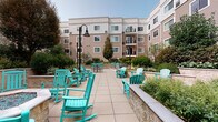 Located at 1 Glover Ave in Norwalk, Connecticut, this well-appointed outdoor courtyard serves as a central gathering space within a multi-story residential building. The patio is finished with large-format tile pavers that provide a clean, durable surface throughout the space. A standout feature is the octagonal fire pit, framed in stone with blue glass accents, and surrounded by vibrant teal Adirondack-style rocking chairs that lend a relaxed, welcoming atmosphere. Additional seating areas with tables and chairs are positioned throughout the courtyard, offering a variety of spaces for both social gatherings and quiet relaxation. The courtyard is bordered by raised stone planters with smooth coping details, filled with an abundance of carefully maintained, diverse plantings including ornamental shrubs, flowering perennials, and mature trees that provide privacy and greenery throughout the seasons. Classic black lamp posts add architectural interest and ensure the space is well-lit. The surrounding multi-story building with balconies wraps around the courtyard, creating an enclosed, sheltered feel while still allowing ample natural light. The overall ambiance is inviting and resort-like, designed for outdoor enjoyment in a well-maintained community setting.