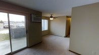 This single-floor space offers a practical and functional layout designed for comfortable everyday living. The main living area is finished with wall-to-wall carpeting that adds warmth underfoot, while a ceiling fan with an integrated light fixture provides both air circulation and ambient lighting. Climate control is well-equipped with a wall-mounted air conditioning unit and baseboard heating running along the perimeter, ensuring year-round comfort regardless of the season. A sliding glass door on one side of the living area allows for natural light to enter the space and provides convenient access to an exterior area. An additional window further brightens the room, contributing to an open and airy feel. The walls feature a neutral, understated color palette, complementing the light-toned carpet and creating a clean, versatile backdrop. A textured ceiling adds a subtle architectural detail throughout. The layout opens toward an adjoining hallway or additional room, suggesting a connected flow between living spaces. Overall, the space presents a straightforward, functional design with essential amenities in place, making it well-suited for a variety of living arrangements.