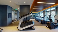 The Laureat Fitness Center in Fort Lauderdale, Florida is a thoughtfully designed, single-level workout facility that combines high-end aesthetics with a comprehensive range of exercise equipment. The space features a striking contemporary interior defined by deep blue-gray accent walls, warm wood-paneled ceiling details, and recessed linear lighting that casts an energizing ambient glow throughout.

Large floor-to-ceiling windows line one side of the facility, inviting abundant natural light and offering views of the surrounding palm-lined Fort Lauderdale streetscape. Sheer drapery softens the light while maintaining an open, airy atmosphere.

The cardio zone is prominently featured, housing a row of treadmills, elliptical trainers, and stair climbers equipped with personal display screens. Strength and resistance training equipment is distributed throughout the space, including cable machines, a leg press, and additional cable-based stations. A glass-walled room provides a semi-private area for focused training, adding functional flexibility to the layout.

Wall-mounted flat-screen displays and built-in audio speakers enhance the workout environment. The flooring transitions between neutral carpet and rubberized fitness surfaces, providing both comfort and practicality. The overall ambiance is polished and modern, reflecting the upscale character of the Laureat property.