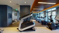 The Laureat Fitness Center in Fort Lauderdale, Florida is a thoughtfully designed, single-level workout facility that combines high-end aesthetics with a comprehensive range of exercise equipment. The space features a striking contemporary interior defined by deep blue-gray accent walls, warm wood-paneled ceiling details, and recessed linear lighting that casts an energizing ambient glow throughout.

Large floor-to-ceiling windows line one side of the facility, inviting abundant natural light and offering views of the surrounding palm-lined Fort Lauderdale streetscape. Sheer drapery softens the light while maintaining an open, airy atmosphere.

The cardio zone is prominently featured, housing a row of treadmills, elliptical trainers, and stair climbers equipped with personal display screens. Strength and resistance training equipment is distributed throughout the space, including cable machines, a leg press, and additional cable-based stations. A glass-walled room provides a semi-private area for focused training, adding functional flexibility to the layout.

Wall-mounted flat-screen displays and built-in audio speakers enhance the workout environment. The flooring transitions between neutral carpet and rubberized fitness surfaces, providing both comfort and practicality. The overall ambiance is polished and modern, reflecting the upscale character of the Laureat property.