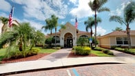 This single-story building showcases classic Florida Mediterranean-style architecture, characterized by a warm cream stucco exterior and a distinctive gabled entryway accented with a decorative arch. The main entrance is framed by elegant arched windows and flanked by wall-mounted lantern-style light fixtures, lending a refined, formal appearance. A paved walkway leads from the street directly to the front entrance, complemented by brick-patterned detailing near the parking area. The building is ADA-accessible, as indicated by designated signage near the entrance. Lush, manicured tropical landscaping surrounds the structure, featuring tall mature palm trees, neatly trimmed hedges, and mulched garden beds with vibrant yellow and green shrubbery. A prominently displayed American flag adds a civic character to the facade. The red tile-accented roofline and extended single-story wings on either side give the building a broad, grounded presence. The overall exterior presents a polished, professional, and welcoming appearance consistent with a community or institutional facility set in a sunny Florida environment.