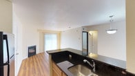 This single-floor residence offers an open and functional layout that seamlessly connects the kitchen and living areas. The kitchen is anchored by rich, dark granite countertops that extend across an L-shaped peninsula, providing generous prep space and a natural divider between the cooking and living zones. Warm honey-toned cabinetry below complements the countertops, and a stainless steel sink with a classic faucet adds a practical finishing touch.

The living area features warm hardwood flooring that runs throughout, contributing to a cohesive and inviting atmosphere. A fireplace with a sleek, dark surround serves as a focal point along one wall, adding both warmth and character to the space. Vertical blinds cover a large sliding glass door or window, allowing natural light to fill the room while offering privacy control.

Neutral, light-toned walls and a textured white ceiling keep the space feeling bright and airy. A doorway visible toward the rear of the unit leads to additional rooms, with wall-mounted lighting adding a subtle accent. Overall, the residence balances practical design with comfortable, everyday livability.
