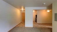 This single-floor space offers a straightforward, functional layout designed for comfortable everyday living. The main living area is finished with wall-to-wall carpeting in a neutral tone, providing a warm underfoot feel that extends throughout the common areas. Flush-mount ceiling light fixtures illuminate the space with a soft, ambient glow, keeping the overall aesthetic clean and understated.

A central partial wall divides the living space, creating a subtle architectural separation while maintaining an open connection to the adjoining kitchen. The kitchen area features tile-style flooring, offering a practical and easy-to-clean surface that contrasts neatly with the carpeted living zone. Stainless steel appliances, including a refrigerator and range, are visible within the kitchen, lending a modern touch to the otherwise classic interior.

Crisp white walls run throughout the space, complemented by light wood-toned baseboards and door frames that add warmth and continuity. A wood-paneled door visible toward the rear of the unit hints at additional rooms or storage beyond the main living area. The overall ambiance is calm and neutral, offering a versatile canvas well-suited for a variety of furnishing styles.