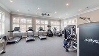 Located at 5150 Jack Byrnes Dr in Murfreesboro, TN, this single-floor fitness center is a comprehensively outfitted facility designed to support a full range of workout routines. The space is bathed in natural light, thanks to an extensive array of large, multi-pane windows with white trim that line multiple walls, offering views of the surrounding community and outdoor pool area. Crown molding and neutral gray walls lend a polished, well-maintained aesthetic throughout.

The cardio section features multiple Life Fitness treadmills equipped with interactive touchscreen consoles, a stair climber, and elliptical machines, all positioned along the windowed walls to take advantage of the natural light and outdoor views. Ceiling fans provide additional air circulation across this area.

A dedicated strength training zone houses a robust selection of Life Fitness cable and resistance machines with blue upholstered seats, catering to a variety of muscle groups. Large wood-framed mirrors are mounted on the walls to assist with form and technique. A separate free weights area includes a full rack of dumbbells, a flat utility bench, and additional seating, complemented by its own set of windows and mirrors.

The carpeted flooring throughout provides comfort underfoot, while recessed lighting ensures the space remains bright and functional at all hours. The overall ambiance is clean, spacious, and inviting.