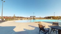 This community amenity center is designed to offer a comprehensive outdoor recreational experience. At the heart of the space is a generously sized swimming pool with a smooth, curved design and gradual entry points, surrounded by an expansive concrete pool deck furnished with lounge chairs and seating arrangements — ideal for relaxing or socializing under open skies. Tall light poles positioned throughout the deck ensure the area remains functional well into the evening hours.

A standout feature is the covered outdoor pavilion, supported by rustic timber-frame posts with a beadboard ceiling. This sheltered patio area connects seamlessly to the main building and houses a striking stone-clad fireplace with a mounted flat-screen television above the mantle, creating a cozy gathering focal point. Curved stone bench seating flanks the fireplace, adding architectural character and functionality.

Adjacent to the pavilion, a dedicated fire pit area features a stone-surround fire pit set into a diamond-shaped concrete pad, providing an additional space for evening gatherings. The perimeter of the facility is enclosed by a black metal fence, with mature trees and surrounding neighborhood views visible beyond. The building itself features light blue and sage green board-and-batten siding, contributing to a welcoming, craftsman-inspired aesthetic throughout the entire amenity complex.