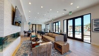 The Parklands Club Room in Ventura, California is a thoughtfully designed community space that seamlessly blends comfort and contemporary style. The open-plan layout is anchored by light hardwood flooring and recessed ceiling lighting that creates a bright, welcoming atmosphere throughout.

The main lounge area features a striking textured stone fireplace wall with a wall-mounted flat-screen TV, complemented by a mix of plush neutral sofas, woven accent chairs, and richly patterned area rugs that add warmth and visual interest. Natural light pours in through an expansive wall of black-framed French doors and floor-to-ceiling windows, offering direct views and access to the outdoor pool area beyond.

A dedicated dining zone accommodates multiple groups with dark-topped tables and teal upholstered chairs arranged on the light wood floors. Glass partition walls provide a sense of separation while maintaining the room's open and airy feel.

A built-in kitchenette area serves as a functional hub, featuring warm wood cabinetry, a stainless steel microwave and appliances, counter seating, and a bold teal tile backsplash with dual wall-mounted screens — ideal for presentations or entertainment. Accent walls in deep teal punctuate the space with color, tying together the overall coastal-modern aesthetic of this versatile club room.