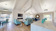 This well-appointed club room in Foster City, California offers an open, single-floor layout that seamlessly integrates a living room, dining area, and kitchen into one cohesive communal space. The most striking architectural feature is the soaring vaulted ceiling with exposed white-painted wood beams and shiplap detailing, which lends the room a bright, airy, and expansive atmosphere. A herringbone-patterned wood-look floor runs throughout, adding warmth and visual texture underfoot.

The living area is anchored by a sleek linear fireplace with a wall-mounted television above, surrounded by multiple seating arrangements featuring teal and blue upholstered chairs and sofas. A statement sputnik chandelier adds a modern accent overhead. Large windows and glass doors on multiple walls flood the space with natural light and provide outdoor views of the surrounding grounds and balconies.

The dining area, set near floor-to-ceiling windows, features a round table with teal upholstered chairs, creating an intimate yet light-filled setting. A decorative sideboard and coastal artwork complete the area.

The kitchen is equipped with stainless steel appliances, taupe shaker cabinetry, a tile backsplash, and a generous marble-topped island with bar seating — ideal for casual gatherings. The overall ambiance is coastal-modern, clean, and welcoming.