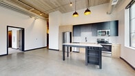 This single-floor space showcases a compelling blend of industrial and contemporary design elements throughout. The architectural character is defined by exposed concrete ceilings with visible ductwork and structural columns, polished concrete floors in the main living areas, and crisp white walls that balance the raw industrial aesthetic.

The open-concept main area centers around a modern kitchen outfitted with dark cabinetry, light wood accents, subway tile backsplash, stainless steel appliances, and a sleek island with pendant lighting suspended from the exposed ceiling above. The generous open floor plan surrounding the kitchen provides flexible living and dining space.

Private rooms feature soft carpeting, exposed concrete columns, and large black-framed windows that frame views of the surrounding urban landscape, including green spaces and city buildings, filling each room with natural light. One area includes direct access to an outdoor balcony through a glass-panel door, further extending the connection to the exterior environment.

A well-appointed bathroom features a soaking tub with tiled surround, matte black fixtures, a wood-toned vanity with white countertop, and large-format tile flooring. Throughout the unit, black baseboards and door trims provide a cohesive, polished contrast to the lighter surfaces, reinforcing the space's modern industrial identity.