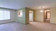 This single-floor residence offers a welcoming and functional layout designed for comfortable everyday living. The main living area is finished with plush wall-to-wall carpeting in a warm beige tone, creating a soft and inviting atmosphere. A standout architectural feature is the pass-through window connecting the living space to the kitchen, allowing for natural interaction between the two areas while maintaining a sense of separation. The kitchen is equipped with wooden cabinetry, a white refrigerator, and tile flooring, providing a practical and clean workspace. A doorway opening directly into the kitchen offers convenient full access. The accent walls are finished in a muted sage green, adding a subtle pop of color that contrasts nicely with the neutral ceiling and trim. A pendant light fixture hangs in the dining area, offering focused overhead lighting and a touch of understated style. Ventilation is supported by ceiling-mounted HVAC vents, ensuring year-round comfort. The overall ambiance is clean, functional, and move-in ready, with a straightforward design that makes excellent use of the available space.