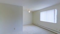 This single-floor space presents a clean and functional layout. The main living area is finished with wall-to-wall carpet in a neutral tone, contributing to a quiet and comfortable atmosphere. Smooth, light-colored walls keep the interior feeling bright and open. A ceiling fan with an integrated light fixture is centrally positioned, providing both air circulation and ambient lighting. A large window fitted with vertical blinds allows natural light to fill the room while offering privacy and light control. Along the base of the exterior wall, a baseboard heater provides supplemental warmth, a practical feature suited for cooler climates. The standard ceiling height and simple, unadorned finishes give the space a straightforward, move-in-ready character. The overall design is minimal and versatile, offering a blank canvas easily adapted to a variety of furniture arrangements and personal styles.