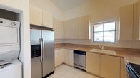 This single-story home presents a clean, light-filled interior with a consistent neutral color palette and a range of practical features throughout. The kitchen is well-equipped with stainless steel appliances, including a side-by-side refrigerator with an ice and water dispenser, a smooth-top electric range, an over-the-range microwave, and a built-in dishwasher. Cream-toned cabinetry provides generous upper and lower storage, complemented by tan countertops and light tile flooring. A window above the sink allows natural light into the space. An arched doorway connects the kitchen to the main living area, which features notably high vaulted ceilings, pendant lighting, and large tile floors that enhance the sense of openness. The living space also includes a louvered closet and additional arched architectural detailing. Bedrooms feature soft carpet flooring, recessed lighting, and windows that bring in natural light and outdoor views. One bedroom includes a bay-style window arrangement. The bathroom features a full vanity with warm wood cabinetry, a large mirror, and a separate water closet housing a tub-shower combination with sliding glass doors and tile surround.