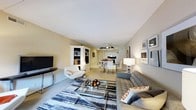 This single-floor residence offers an open-concept layout that integrates the living, dining, and kitchen areas into one cohesive and functional space. The living area is anchored by a plush gray sectional sofa accented with decorative orange-and-white pillows, complemented by a sleek glass-and-chrome coffee table and a patterned area rug in navy and gray tones. A white accent chair and a built-in shelving unit housing a flat-screen television add both comfort and practicality to the space. The walls are finished in warm neutral tones, contributing to an inviting and relaxed ambiance throughout. A vaulted ceiling with subtle angled lines adds architectural interest and enhances the sense of openness. The dining area, positioned centrally, features a glass-top table with modern chairs, bathed in soft light from a ceiling fan overhead. The kitchen beyond is finished in white cabinetry with clean lines, providing ample storage and a bright, airy feel. Framed artwork adorns the walls, adding a curated, personal touch. The overall design balances contemporary style with everyday livability, making this a well-appointed and thoughtfully arranged single-level home.