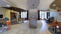 This thoughtfully designed single-floor space offers an open-concept layout that seamlessly blends functionality with contemporary style. At its heart is a large, curved reception or conference table crafted in warm wood tones, serving as the visual anchor of the room. A striking stone-clad fireplace rises behind it, adding a refined focal point to the space.

The ceiling design is a standout architectural feature, combining a dramatic oversized circular pendant fixture with a slatted wood panel installation, adding depth and texture overhead. Pendant lights are suspended at varying heights throughout, contributing to an inviting, layered ambiance.

Natural light pours in through large windows dressed with rich, floor-length curtains, framing views of the outdoors and creating a bright, welcoming atmosphere. The flooring transitions between a patterned area rug in the central lounge area and warm hardwood, helping to define distinct functional zones.

To one side, a café-style bar counter with high stools provides a casual gathering spot alongside a coffee station. On the opposite end, a dedicated workstation area with desks and ergonomic chairs offers a productive environment. Exposed brick accents add an industrial warmth that balances the polished, modern finishes throughout the space.