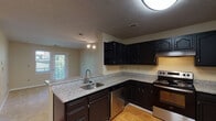 This single-floor space is thoughtfully laid out to maximize functionality across its key areas. The kitchen serves as a central highlight, featuring dark espresso-toned cabinetry with ample upper and lower storage, granite-look countertops, and stainless steel appliances including an electric range and dishwasher. A peninsula with an undermount sink provides additional workspace and separates the kitchen from the adjoining living area. Light hardwood-style flooring runs throughout the kitchen, adding warmth to the space.

The living area is carpeted and benefits from natural light through a glass door and adjacent window that provide views of the surrounding greenery and direct outdoor access. Track lighting adds a practical and modern touch to the open layout.

The bedroom is generously proportioned with plush carpeting, neutral walls, and a sizable window fitted with blinds, allowing for controlled natural light. A textured ceiling is consistent throughout the unit.

The bathroom is well-appointed with a dark-toned vanity cabinet, a single-basin sink with a large mirror above, and a full-size soaking tub and shower combination. Light hardwood-style flooring continues here, tying the space together cohesively. Overall, the unit offers a clean, neutral aesthetic with practical finishes throughout.