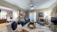 This single-floor residence offers a cohesive and elegantly curated interior, blending contemporary design with warm, inviting finishes throughout. The entry hallway, lined with rich hardwood flooring, sets the tone as it opens to the main living areas adorned with bold abstract artwork in gold-framed displays. Crown molding runs throughout, adding a refined architectural detail to the neutral-toned walls. The living room is a standout space, featuring a ceiling fan, vertical blind-covered sliding glass door, and a sophisticated arrangement that balances comfort with style. The dining area includes a mirrored wall accent that amplifies light and space, complemented by hardwood floors and crown molding. The kitchen is functional and well-equipped, featuring light maple cabinetry, light stone countertops, white appliances, and tile flooring. The bedroom is a serene retreat with plush carpet, large windows with drape treatments, and crown molding, while the walk-in closet provides generous storage with dual hanging rods. A full bathroom offers a tub-shower combination, tile flooring, and a wood-toned vanity. The in-unit laundry area with tile flooring adds everyday convenience to this well-appointed residence.
