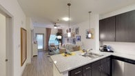 This single-floor residence showcases an open-concept layout that seamlessly connects the kitchen and living areas, creating a spacious and functional environment. The kitchen is anchored by a generous granite-topped island with an integrated sink, complemented by dark espresso cabinetry with sleek hardware. Pendant lights hang above the island, providing both task lighting and a stylish design element.

The transition into the living area is effortless, where light hardwood-style flooring runs throughout, enhancing the sense of continuity. A ceiling fan with a central light fixture serves the living space, contributing to both comfort and illumination. Large windows allow abundant natural light to fill the room, creating a warm and welcoming ambiance, while a glass door provides access to an outdoor area.

The living space is well-proportioned, offering ample room for comfortable furnishings. White walls serve as a neutral backdrop, highlighted by a curated gallery wall arrangement. The overall design balances modern finishes with a bright, airy aesthetic, making the space feel open and inviting throughout.