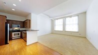 This single-floor space offers a well-designed open-plan layout that combines the living area and kitchen into one cohesive environment. The living area is finished with warm hardwood flooring that transitions into plush wall-to-wall carpeting, creating a comfortable and inviting atmosphere. A row of generously sized windows along one wall allows abundant natural light to pour in, offering views of the surrounding urban streetscape and contributing to the bright, airy feel of the space. The clean white walls and light-toned ceiling enhance the sense of openness throughout. The kitchen is positioned to one side and features dark wood cabinetry, a dark countertop, and a breakfast bar that provides a natural boundary between the cooking and living areas — ideal for casual dining or entertaining. Structural beams are subtly visible along the ceiling, adding an architectural character that hints at the building's heritage. The overall ambiance is clean, modern, and functional, making the most of the available space through thoughtful layout and natural light. The combination of hardwood and carpet flooring, along with the open design, creates a versatile living environment suited to a variety of lifestyles.