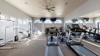 This fitness center offers a thoughtfully designed workout space spread across a single floor, providing ample room for a comprehensive range of exercise equipment. The space features soaring vaulted ceilings that lend an open, airy feel, complemented by a large ceiling fan that promotes airflow throughout. Arched transom windows line the upper walls, flooding the interior with natural light while maintaining privacy at eye level, and recessed lighting ensures consistent illumination throughout the day.

The workout floor is carpeted and organized to accommodate a diverse selection of equipment. Cardio stations are prominently featured, including multiple treadmills, elliptical trainers, and a stationary bike, several of which are equipped with personal display screens. A dedicated strength training area houses cable and pulley machines, a multi-function weight station, and benches suited for a variety of exercises.

Full-length mirrors span one wall, visually expanding the space and allowing users to monitor their form during workouts. The neutral gray walls and clean white trim contribute to a modern, polished aesthetic. A water vending machine is conveniently accessible near the entry. The overall ambiance is bright, functional, and welcoming, making this fitness center a practical and well-appointed facility for a full-body workout routine.