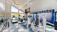 Located at 8114 W Britton Rd in Oklahoma City, Oklahoma, this fitness center offers a thoughtfully designed exercise space filled with an impressive array of equipment and modern amenities. The room is defined by its striking vaulted, cathedral-style ceilings that soar overhead, creating an open and energizing atmosphere. Large, floor-to-ceiling angled windows along one wall flood the space with abundant natural light while offering views of an outdoor pool area, complete with yellow umbrellas, adding a vibrant backdrop to workouts. A ceiling fan and wall-mounted flat-screen television enhance comfort and entertainment during exercise sessions.

The flooring features a bold geometric hexagonal tile pattern in shades of blue, gray, and white, lending the space a contemporary, polished aesthetic. Equipment includes Life Fitness cardio machines such as treadmills, ellipticals, and a stationary bike, along with multiple strength training machines and a free-weight bench. A full dumbbell rack is positioned in front of a large wall mirror, maximizing functional use of the space.

A dedicated functional training zone features a bold blue multi-purpose rig with monkey bars, suspension straps, and resistance band attachments, set over an artificial turf mat with battle ropes — ideal for dynamic, high-intensity workouts. Large sliding glass doors provide easy access and additional natural light, completing this well-rounded and inviting fitness facility.